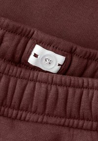 Tissu en coton bordeaux avec une texture côtelée. Comprend une étiquette blanche avec un trou de bouton et un bouton transparent sur la ceinture.