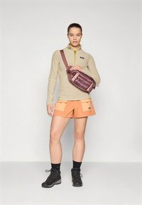 Tilsittet fleecegenser i tan med glidelåskrage, kombinert med oransje shorts med lommer og svarte tursko. En brun skulderveske.