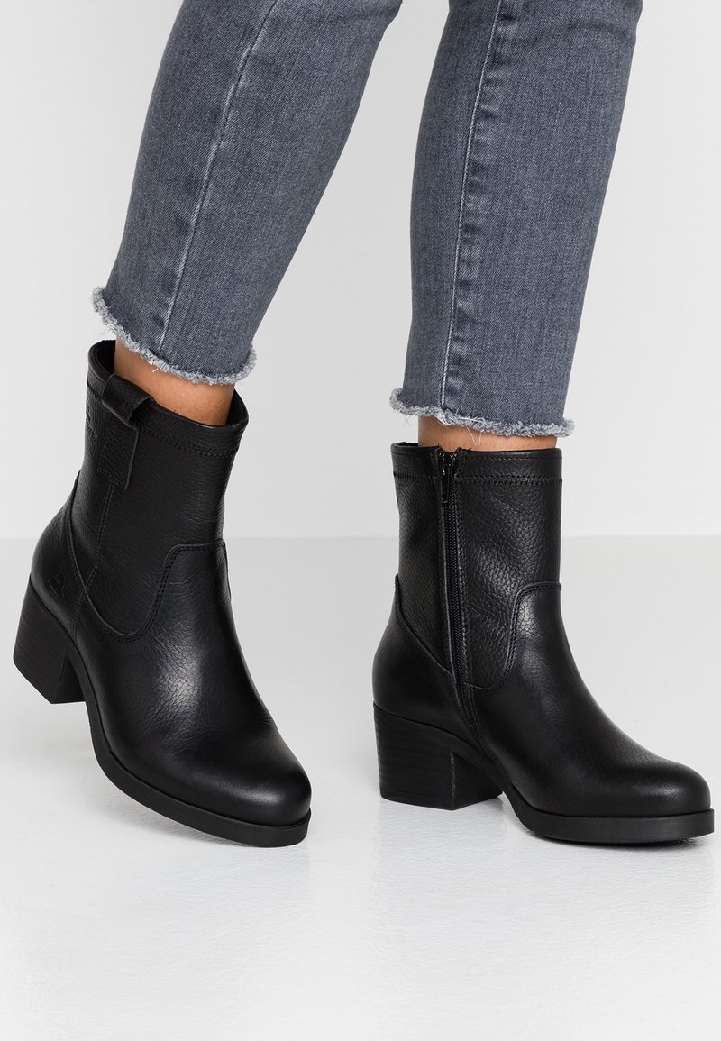 Bottes cheville en cuir noir avec talons carrés, portées avec un jean gris à ourlet effiloché, présentées sur un sol et un fond blancs.