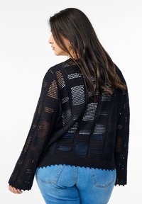 Svart stickad cardigan med ett öppet vävmönster, långa ärmar och scalloped nederkant. Texturerad med kontrasterande rutmönster.