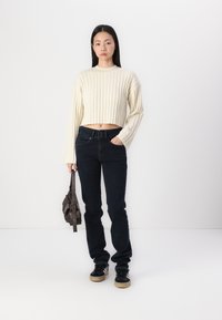 Kremfarget ribbestrikket cropped genser, mørkeblå jeans, svarte sneakers med brune såler, holder en brun veske. Enkelt design, avslappet passform.