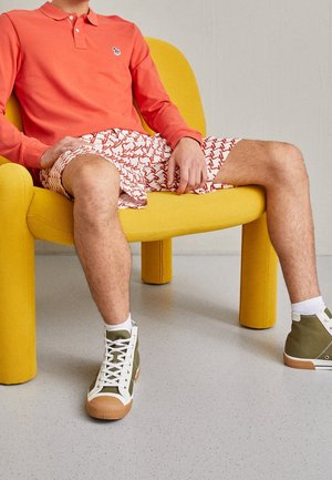 Mand iført koralfarvet langærmet polo, mønstrede shorts og grønne høje sneakers sidder i en gul moderne lænestol.