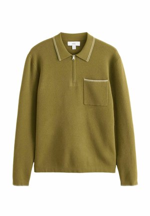 Man in green long-sleeve polo shirt and black pants leaning against a white wall on a tiled outdoor patio.