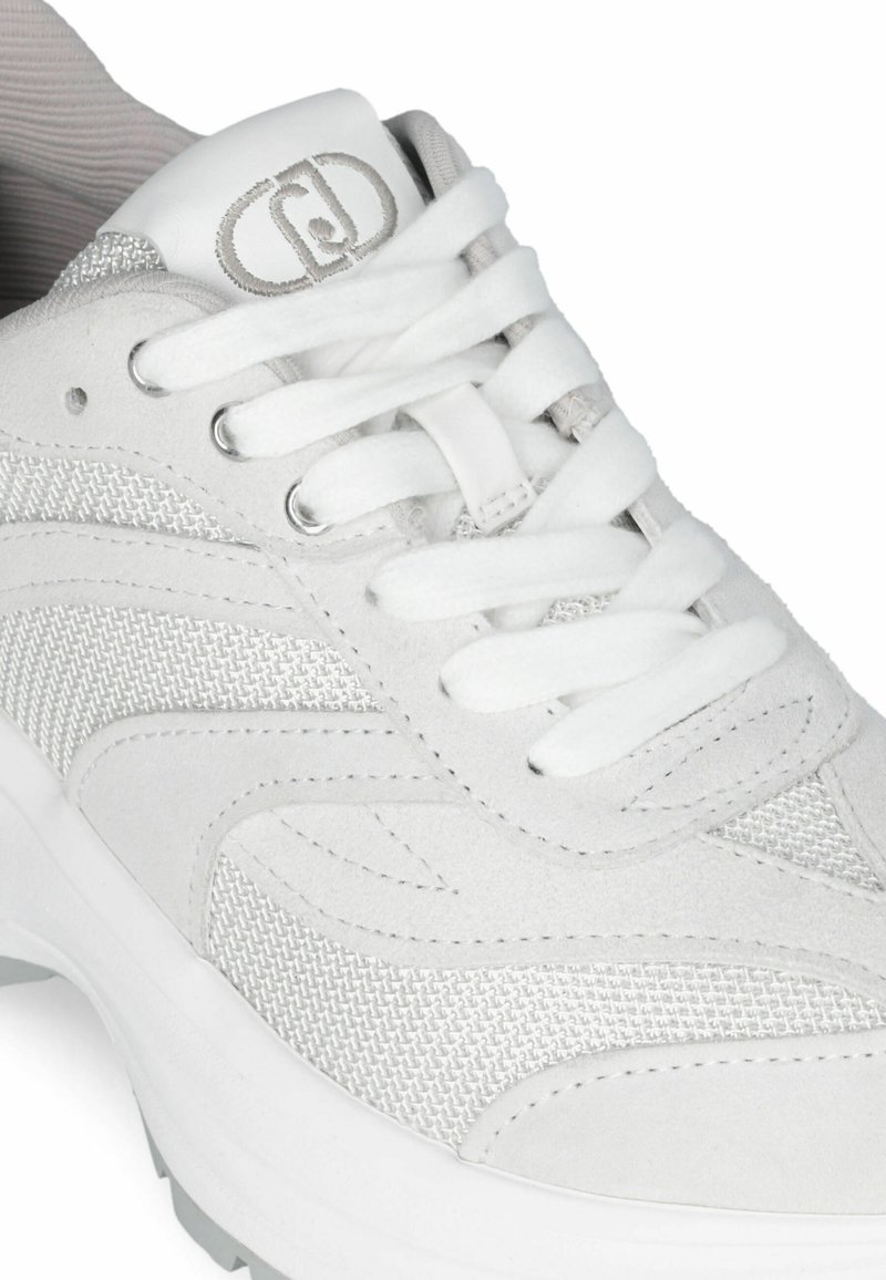 Zapatilla blanca con materiales de ante y malla, punta redonda, cordones, logo en la lengüeta, con una suela blanca gruesa y acentos texturizados.