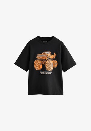 Melnā īsām piedurknēm T-kreklā ar oranžu monstru kravas automašīnas grafiku un tekstu "Monstru Mānija, Giants mājas" uz priekšpuses.