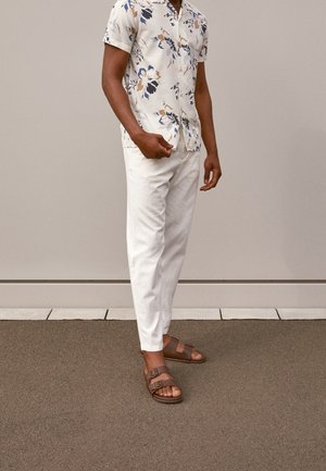 Mand iført beige blomsterprint kortærmet skjorte, hvide bukser og brune sandaler med spænde, stående på fortov foran en grå væg.