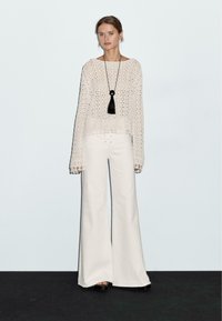 Hellbeige gestrickter Pullover mit weiten Ärmeln über weißen schmalen Hosen. Auffällige schwarze Quasten-Halskette; eleganter, strukturierter Stoff.
