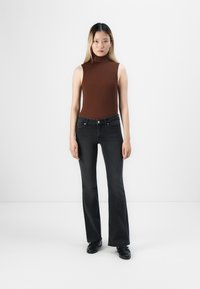 Jonge vrouw staat, gekleed in een mouwloos bruin coltrui-top, zwarte bootcut jeans en zwarte schoenen tegen een effen witte achtergrond.