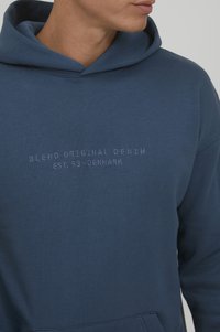 Marineblaue Kapuzenjacke aus weichem Baumwollmaterial mit einer vorderen Kängurutasche. Mit aufgesticktem Text: "BLEND ORIGINAL DENIM EST. 93-DANEMARK."