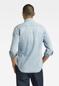 Light blue denim shirt with rolled cuffs, a pointed collar, and a curved hem. The fabric has a soft, textured finish with subtle stitching details.
