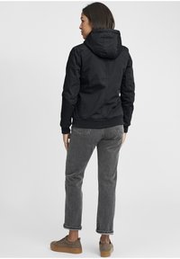 Black hooded jacket with a fitted design, elastic cuffs, and a ribbed waistband, paired with gray jeans and textured brown shoes.