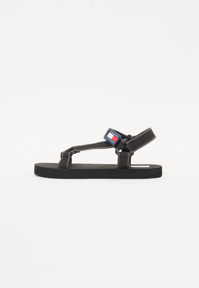 Black flat sandals with fabric straps and a textured sole. Features a logo patch in red, white, and blue on the ankle strap.