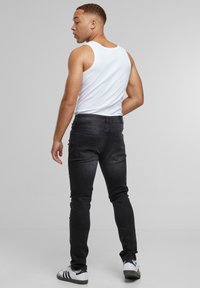 Mees valges tank-topis, mustades slim-joonistes teksades ja valgetes tossudes mustade ribadega, seismas ja üle õla vaatamas halli tausta taustal.