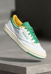 Biele kožené tenisky so zelenými akcentmi a žltým podšívaním. Majú štruktúrovanú gumovú podrážku a výrazné logo Reebok na boku.