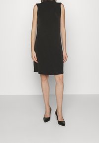 Svart ärmlös klänning med hög halsringning och sidoklyfta, tillverkad av ett jämnt tyg. Matchas med svarta högklackade skor.