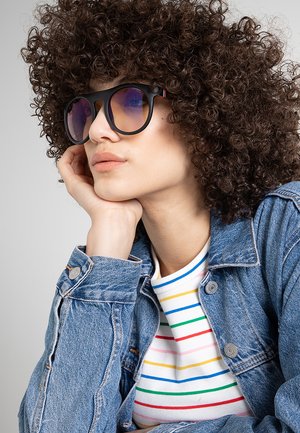 Donna con capelli ricci che indossa occhiali da sole scuri, una maglia a righe e una giacca di jeans blu, appoggiando il viso sulla mano.