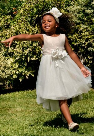 Ragazza giovane in abito bianco e fascia per capelli che balla all'aperto sull'erba con cespugli verdi sullo sfondo in una giornata di sole.