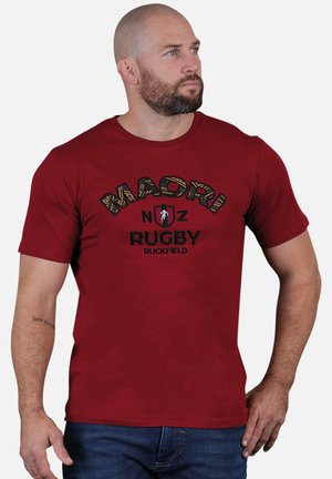T-shirt en coton rouge avec "MAORI NZ RUGBY RUCKFIELD" imprimé en noir et en police jaune texturée. Manches courtes et col rond.
