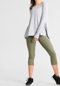 Helles graues Langarmoberteil mit lockerer Passform und asymmetrischem Saum, kombiniert mit olivgrünen Caprileggings. schwarze Sportschuhe runden das Outfit ab.