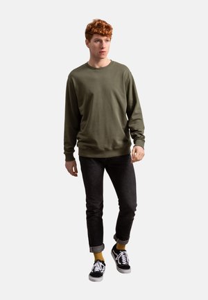 Olive green sweatshirt with long sleeves, round neck, paired with dark jeans and black sneakers, featuring yellow socks. Subtle texture.