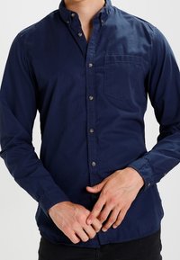 Tummansininen nappipaita, joka on valmistettu puuvillasta, jossa on teräväkauluksellinen malli, pitkät hihat ja rintatasku; sileä kangas ja normaali istuvuus.