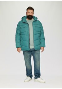 Turkusowa kurtka puchowa z dwoma kieszeniami na piersi i zamkiem błyskawicznym z przodu, połączona z jasnym swetrem i niebieskimi dżinsami; białe trampki dopełniają całość.