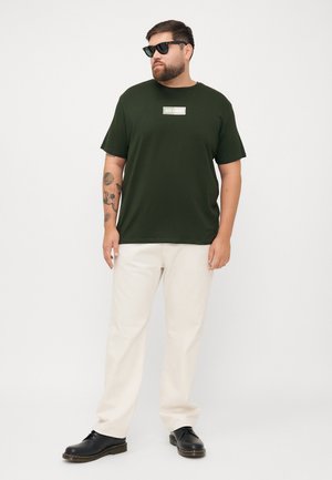 Dunkelgrünes Baumwoll-T-Shirt mit kurzen Ärmeln und einem Etikett auf der Brust; kombiniert mit cremefarbenen Hosen und schwarzen Schuhen.
