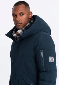 Mörkblå puffjacka med huva, dragkedjeförsedd armficka och texturerad framsida, lager på lager med en rutig svart och vit scarf.