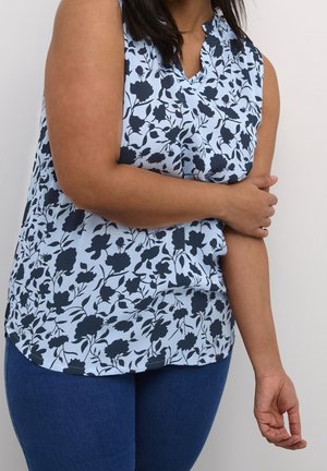 Femme en blouse sans manches bleu clair à motif floral foncé et jean bleu, tenant son bras droit avec la main gauche.