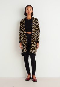 Cardigan s uzorkom leopard kože u crnoj i bež boji, s otvorenim prednjim dijelom, nošen preko crnog kratkog topa i crnih tajica. Crvene cipele s potpeticama.