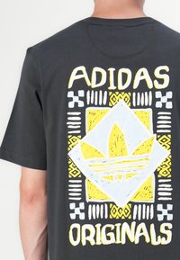 Juodas medvilnės marškinėliai su dideliu geltonu ir baltu grafiniu piešiniu, kuriame yra Adidas logotipas ir tribaliniai raštai nugarinėje pusėje. Trumpomis rankovėmis.