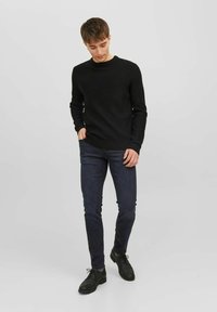 Svart stickad tröja med ribbad textur, rundad hals, kombinerad med mörkblå slim-fit jeans och svarta snörskor. Enfärgad bakgrund.