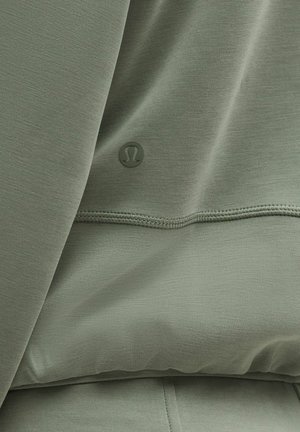 Olivzaļš kapučjakas džemperis ar gludu tekstūru, ar mazu logotipu un redzamu šuvju līniju gar apakšējo malu. Audums ir mīksts un viegls.