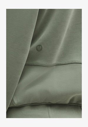 Olivzaļš kapučjakas džemperis ar gludu tekstūru, ar mazu logotipu un redzamu šuvju līniju gar apakšējo malu. Audums ir mīksts un viegls.