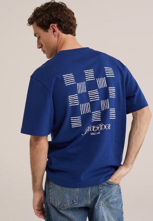 Mann trägt blaues T-Shirt mit weißen abstrakten grafischen Quadraten und dem Schriftzug „BLUE RIDGE SINCE 1986“, kombiniert mit blauen Jeans aus Denim.