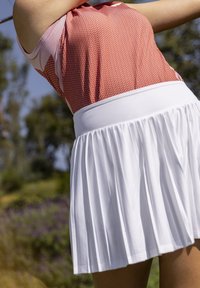 Donna con una gonna plissettata bianca e un top senza maniche a motivo rosso che colpisce una pallina da golf all'aperto.