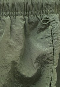 Tkanina olivové barvy s mačkanou strukturou a staženým pasem. Obsahuje šňůrku a detail bočního švu. Lehký design.