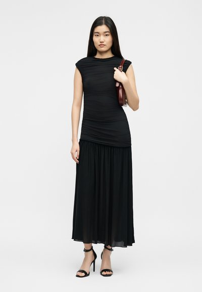 Γυναίκα με μακριά σκούρα μαλλιά, ντυμένη με μαύρο αμάνικο φόρεμα με ρυτίδες, μαύρα σανδάλια με ψηλά τακούνια και κρατώντας καφέ τσάντα ώμου.