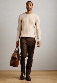 Man walking indoors wearing cream cable knit sweater, dark brown pants, dark loafers, carrying a brown leather duffel bag in left hand.