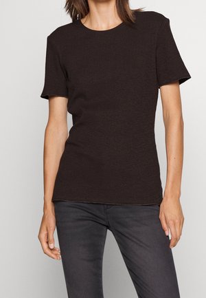 Osoba ubrana w dopasowaną czarną prążkowaną koszulkę z krótkim rękawem i ciemnoszare dżinsy na jednolitym jasnym tle.