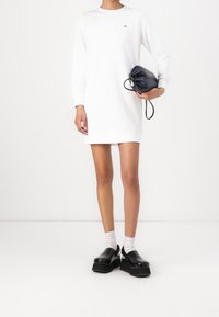 Weißes langärmeliges Kleid mit rundem Ausschnitt, kombiniert mit schwarzen klobigen Slip-on-Schuhen und einer kleinen schwarzen Tasche mit Kordelzug.