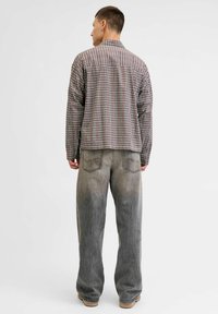 Camicia a quadri multicolore con maniche lunghe, abbinata a jeans grigi larghi e con una vestibilità casual, in piedi su uno sfondo bianco.