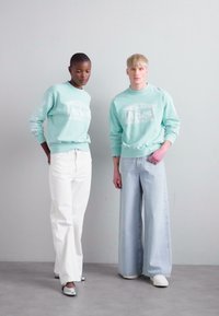 Hanorace de culoare verde mentă cu text imprimat, asortate cu pantaloni albi largi și blugi de culoare albastru deschis. Fondul are o culoare neutră.