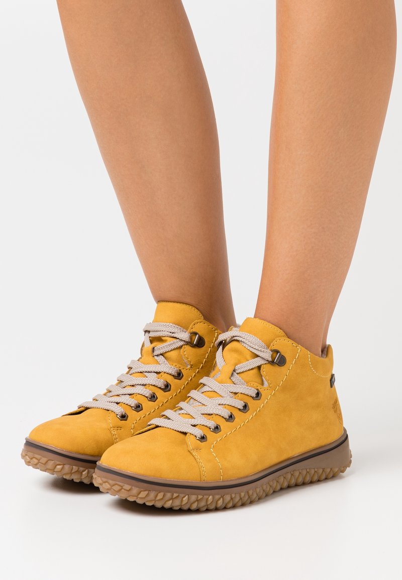 Zapatillas de ante amarillas con cordones beige y suela de goma texturizada, que presentan detalles de costura y un diseño de corte bajo.