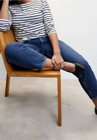 T-shirt rayé à manches longues en blanc et bleu marine, associé à un jean cropped bleu foncé. Assis sur une chaise en bois à la surface lisse. Chaussures plates noires.