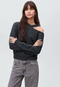 Črn, bleščeč off-shoulder pulover z dolgimi rokavi, v kombinaciji s sivimi jeans hlačami s kovinskimi dodatki, ki prikazujejo teksturiran videz tkanine.