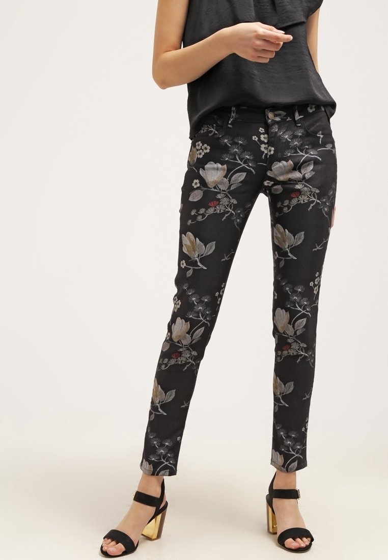 Mujer vistiendo pantalones ajustados negros con estampado floral y tacones abiertos negros con tacones dorados cuadrados, de pie contra un fondo claro y liso.