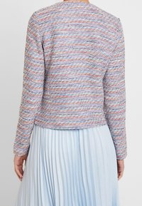 Meer gekleurde geweven jas met een gestructureerde afwerking, ronde halslijn en lange mouwen, gecombineerd met een lichtblauwe geplooide rok.