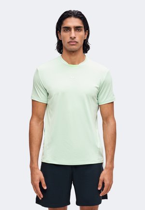 Homme aux cheveux foncés mi-longs portant une chemise vert clair à manches courtes et un short noir, debout devant un fond uni.