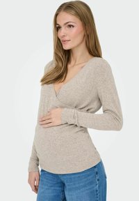 Beżowa bluzka z długim rękawem w stylu wrap z miękkiej tkaniny, z głębokim dekoltem w kształcie litery V i dopasowanym krojem, noszona na niebieskich dżinsach z lekko zaszytym przodem.